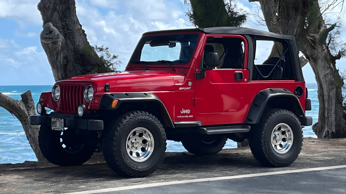 Red Jeep parked by the ocean with trees in the background.