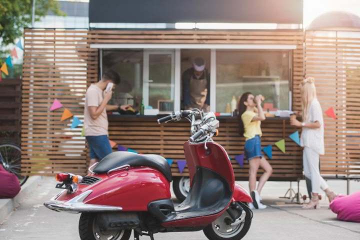 Red scooter parked near a food truck with people ordering and talking.
